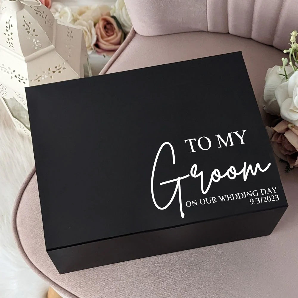 A black gift box with the text 'To My Groom On Our Wedding Day' printed in gold vinyl on the lid, placed on a chair with a floral decoration and a personalized touch.